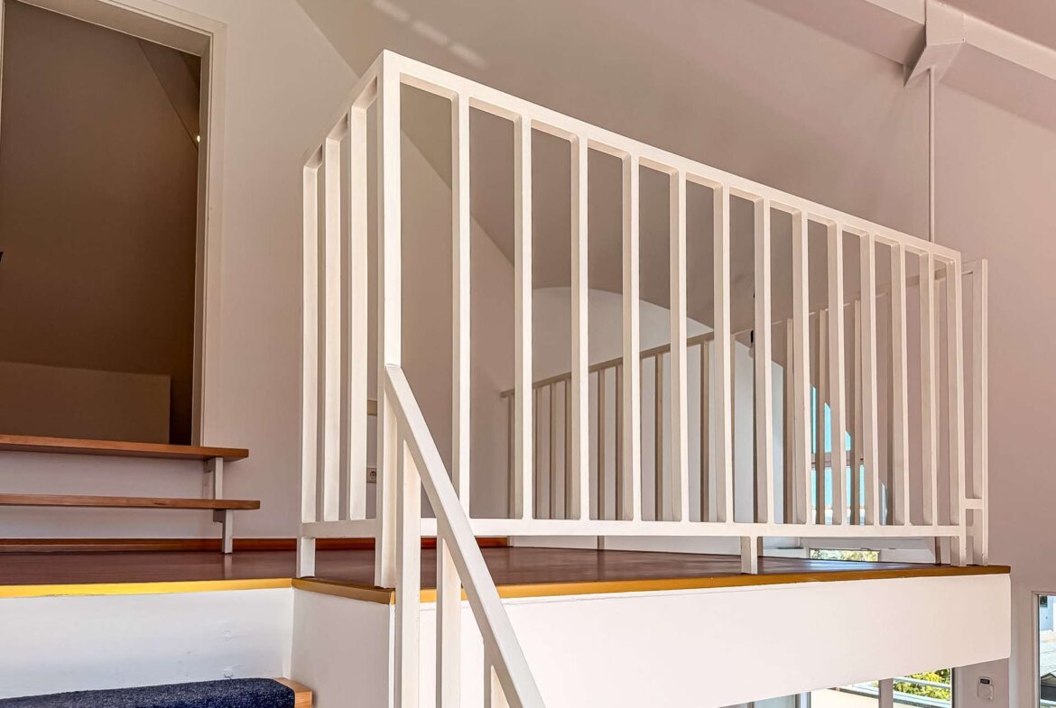 Indoor stairwell with white vertical-bar railing and wood steps, leading to a mezzanine balcony area.