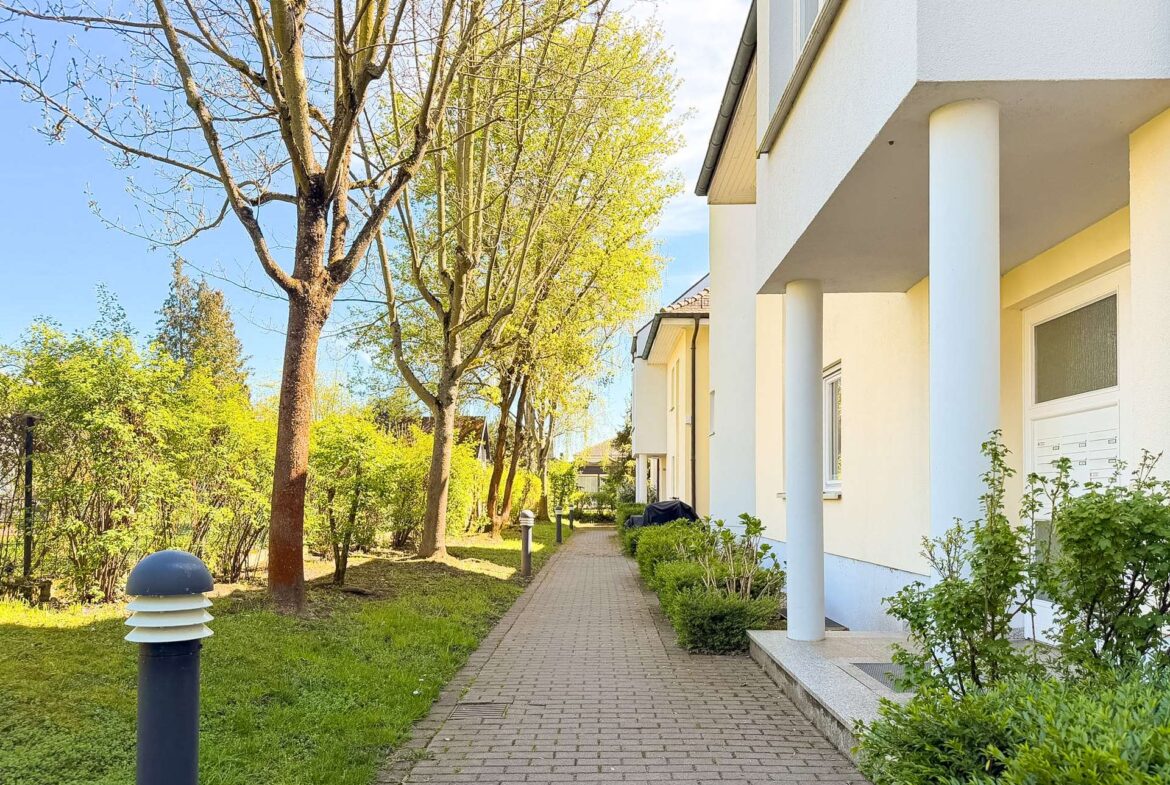 Brick-paved sidewalk beside a white apartment building with blue columns and green shrubs, under a sunny blue sky.