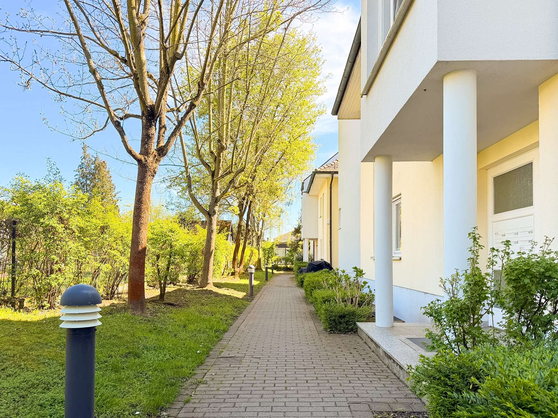 Brick-paved sidewalk beside a white apartment building with blue columns and green shrubs, under a sunny blue sky.