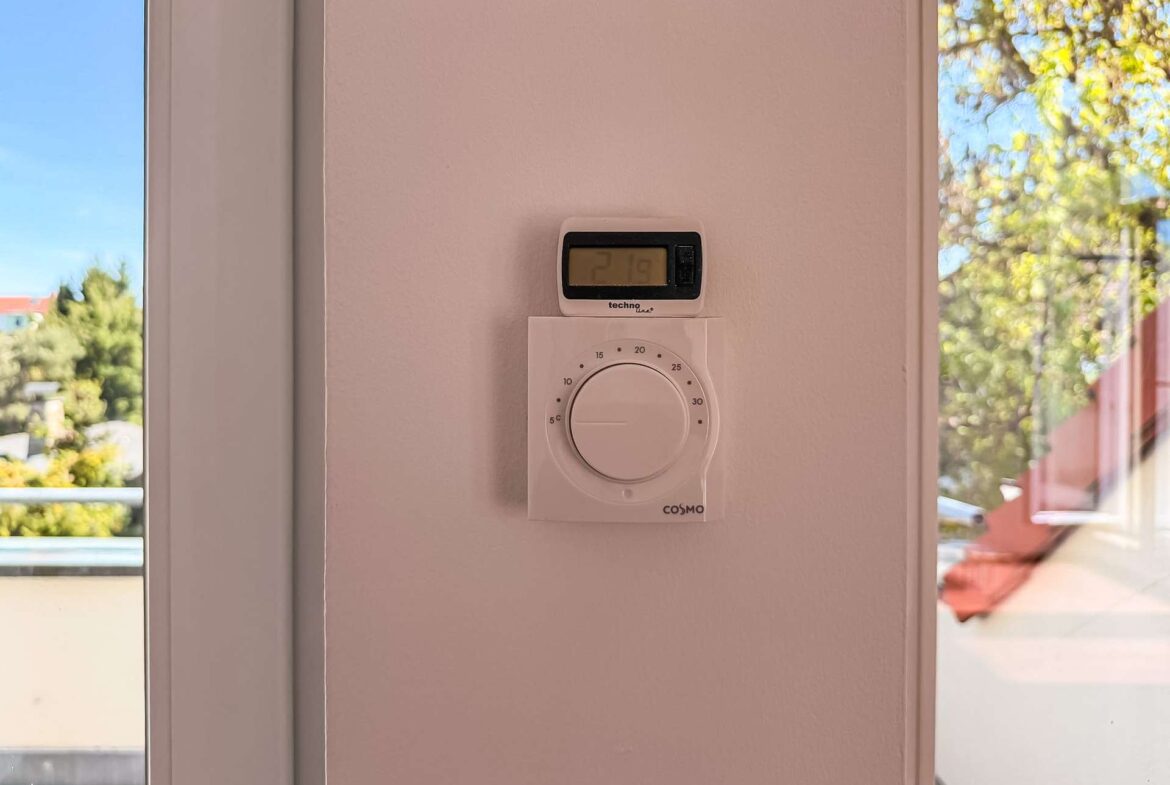 Wall-mounted thermostat with digital display showing 21.9°C and a large circular control knob beneath, on a light pink wall; windows with outdoor trees visible.
