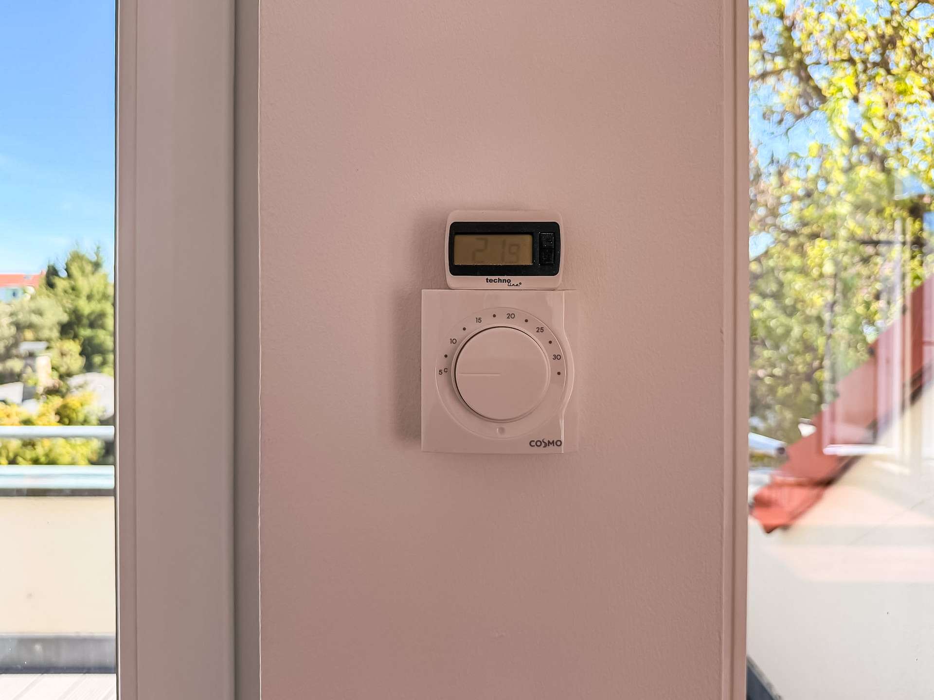 Wall-mounted thermostat with digital display showing 21.9°C and a large circular control knob beneath, on a light pink wall; windows with outdoor trees visible.