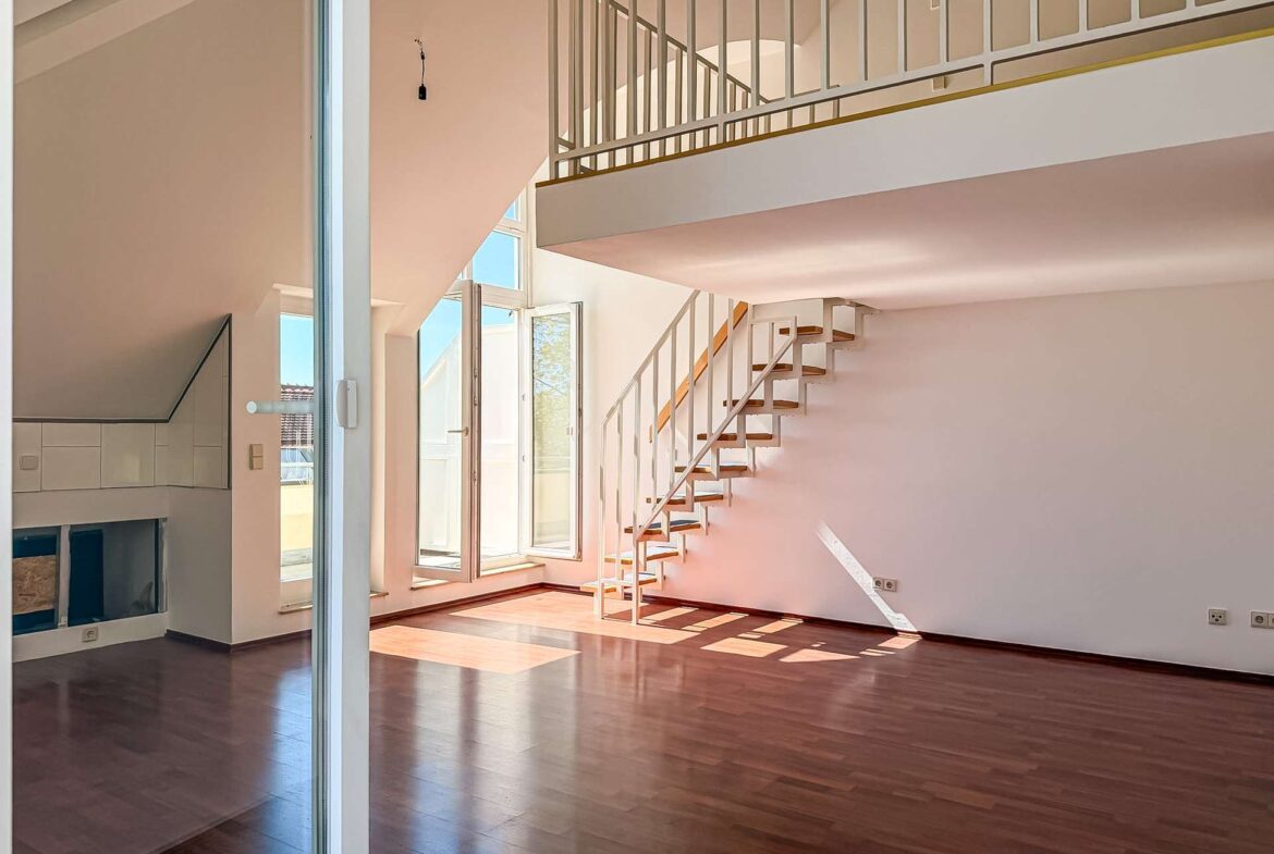 Bright, open living area with dark wood flooring, large windows, and a metal-railed staircase to a mezzanine.
