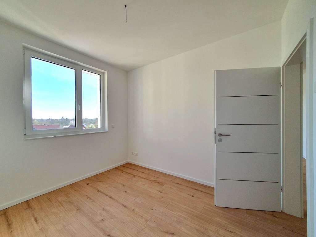 A bright empty room with white walls, a large window, and a light wooden floor; a white door with horizontal grooves is ajar on the right side.