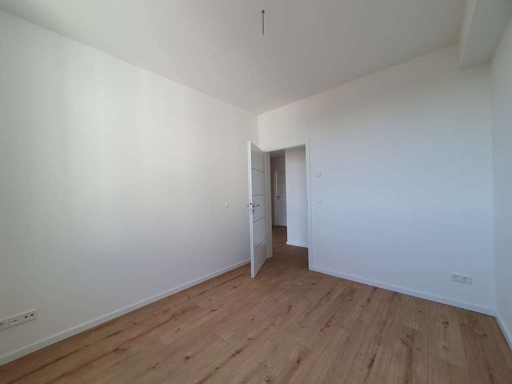 Empty white room with light wood flooring and an open door showing another room beyond.