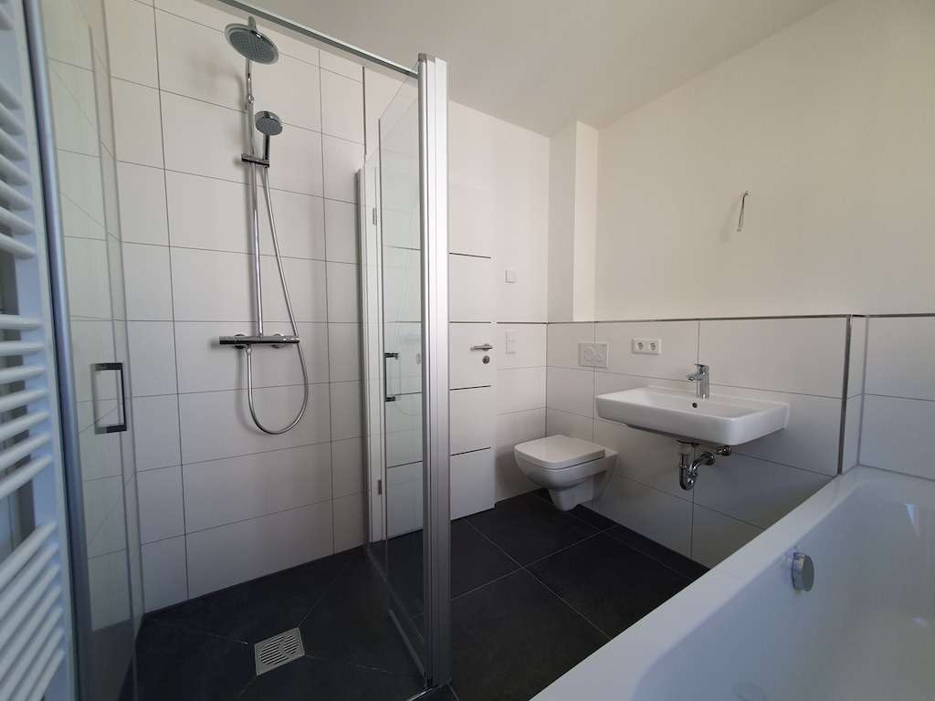 Modern bathroom with a glass-enclosed shower, wall-mounted sink, and wall-hung toilet, white tiles, and a bathtub edge visible to the right