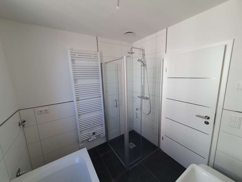 Modern white bathroom with a glass corner shower, towel warmer, white door, and dark floor tiles.