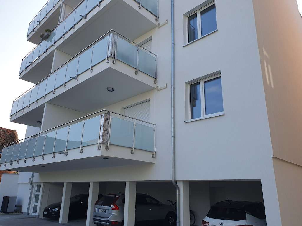 Modern white apartment building with glass-front balconies and a covered ground-floor parking area with cars visible beneath apartments