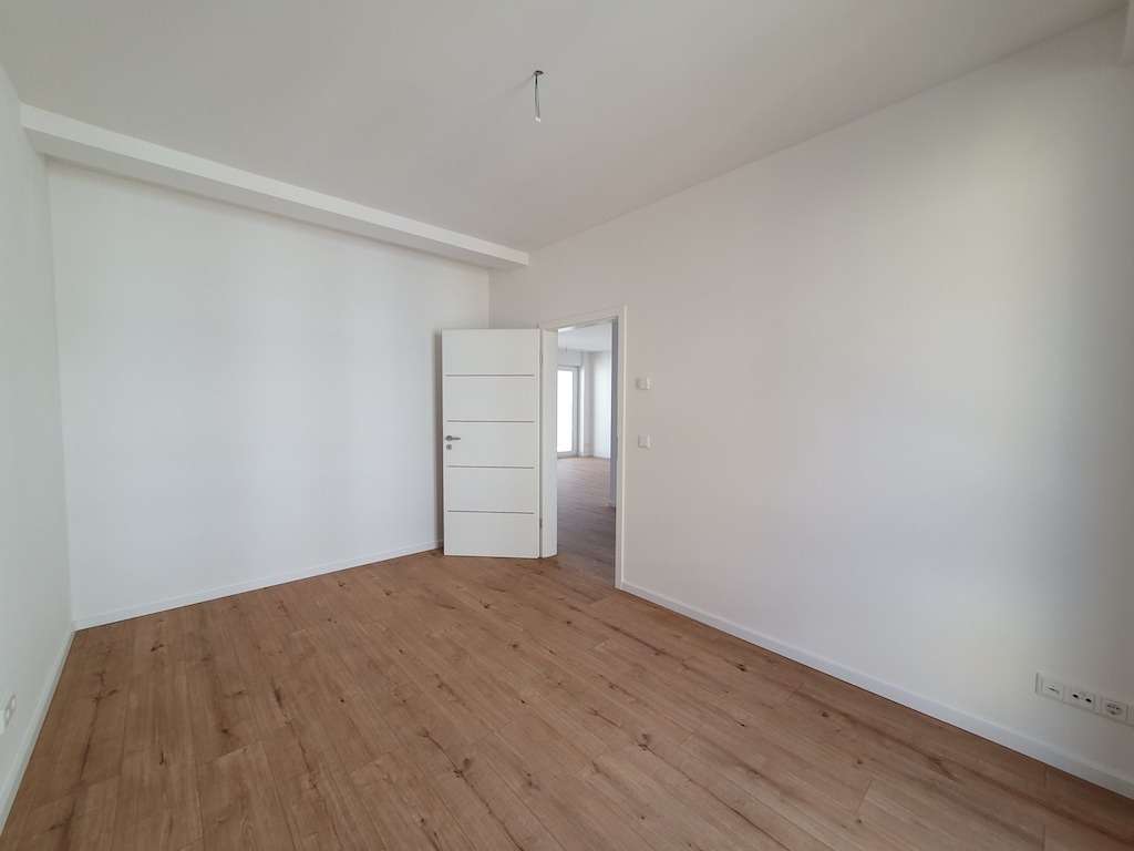 Empty white room with light wood floor and an open door revealing another sunlit room in the next space.