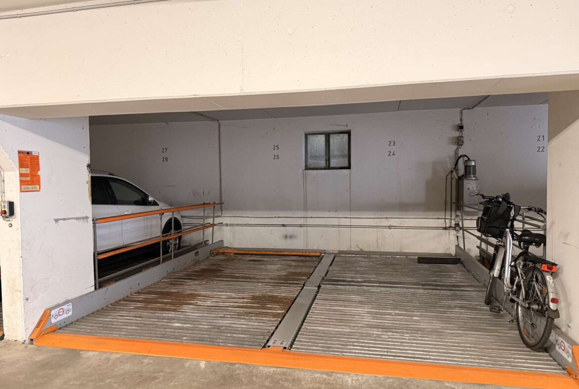 Underground parking area with a car on the left, bicycles on the right, orange safety rails, and numbered wall panels (23–28) near a small window.