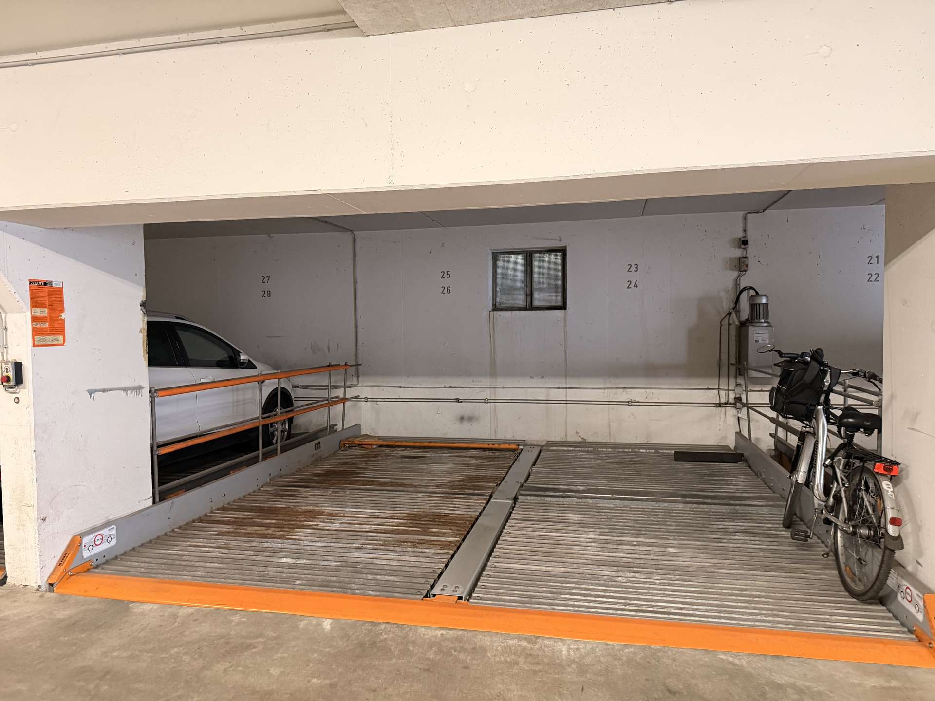 Underground parking area with a car on the left, bicycles on the right, orange safety rails, and numbered wall panels (23–28) near a small window.
