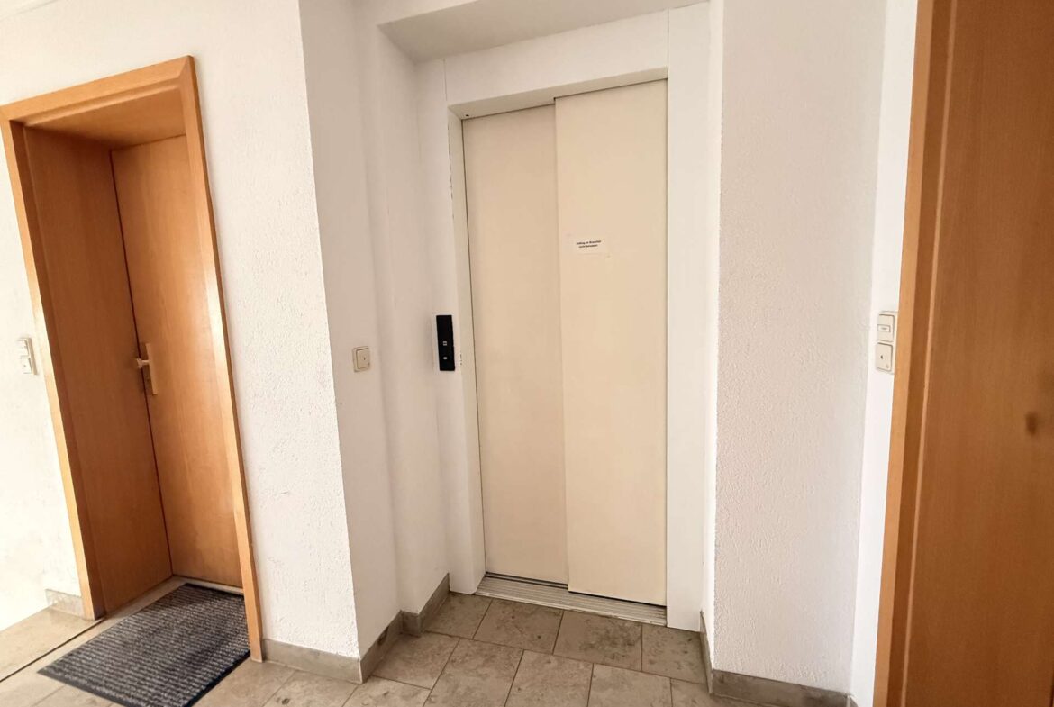 An elevator with closed white doors in a hallway, flanked by wooden doors and a black call button panel on the left.
