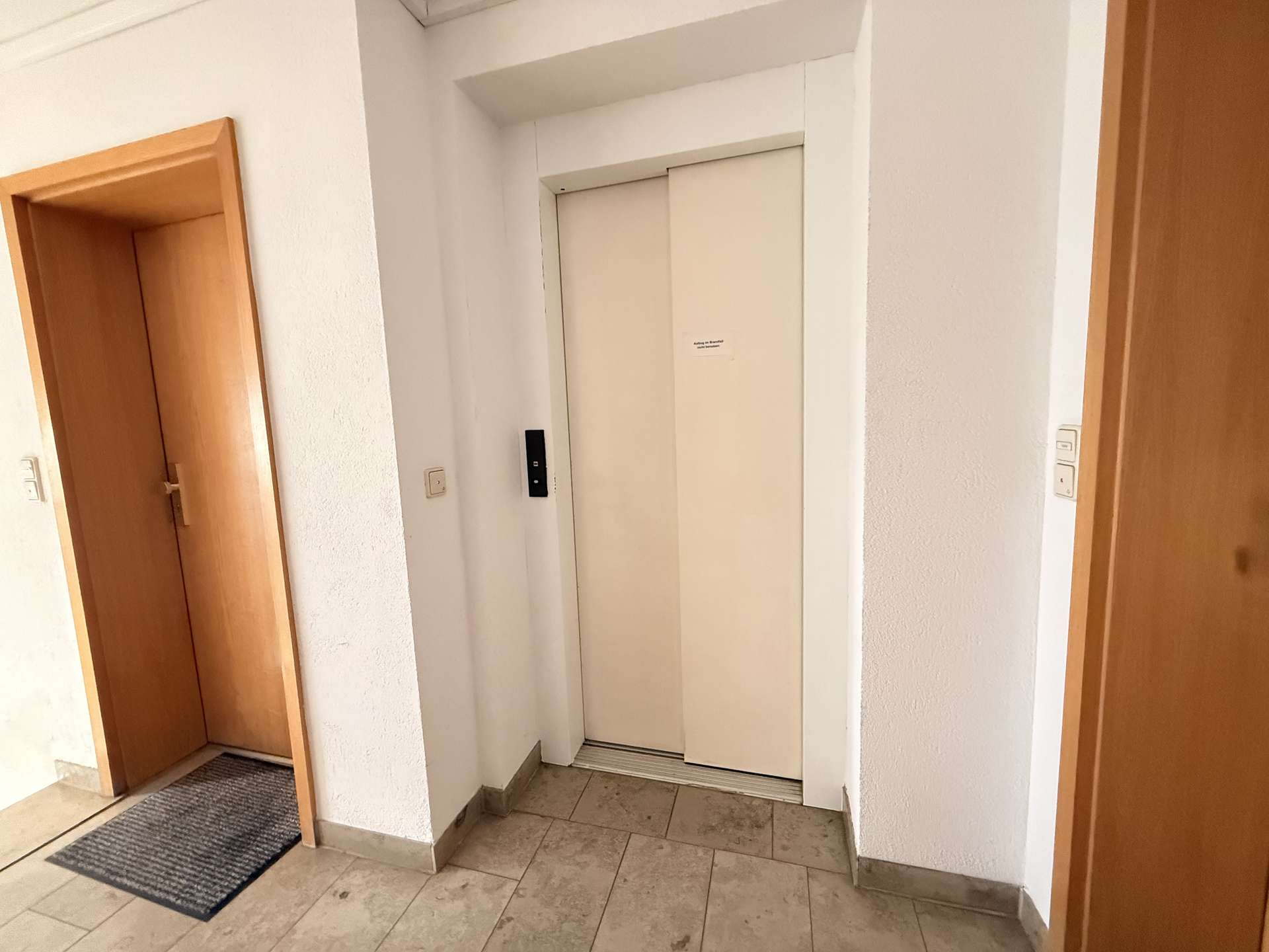 An elevator with closed white doors in a hallway, flanked by wooden doors and a black call button panel on the left.