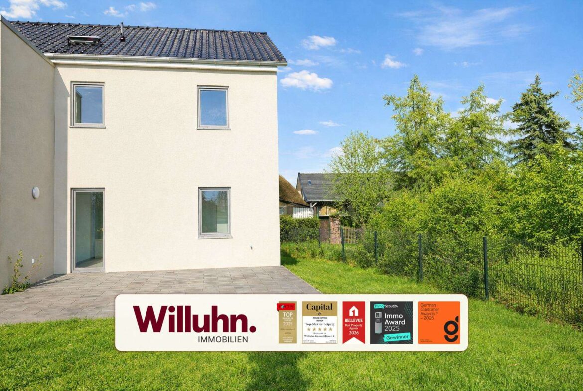 Two-story white house with a tiled roof, a paved patio, green lawn, and a Willuhn Immobilien banner with awards in front.