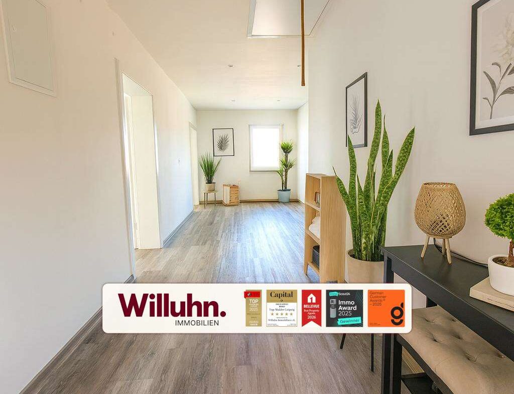 Bright hallway interior with wooden floor, potted plants, framed botanical art, and a wooden console with a wicker lamp; a Willuhn Immobilien banner across the bottom.
