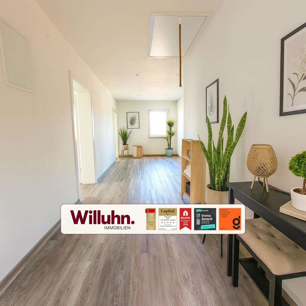 Bright hallway interior with wooden floor, potted plants, framed botanical art, and a wooden console with a wicker lamp; a Willuhn Immobilien banner across the bottom.