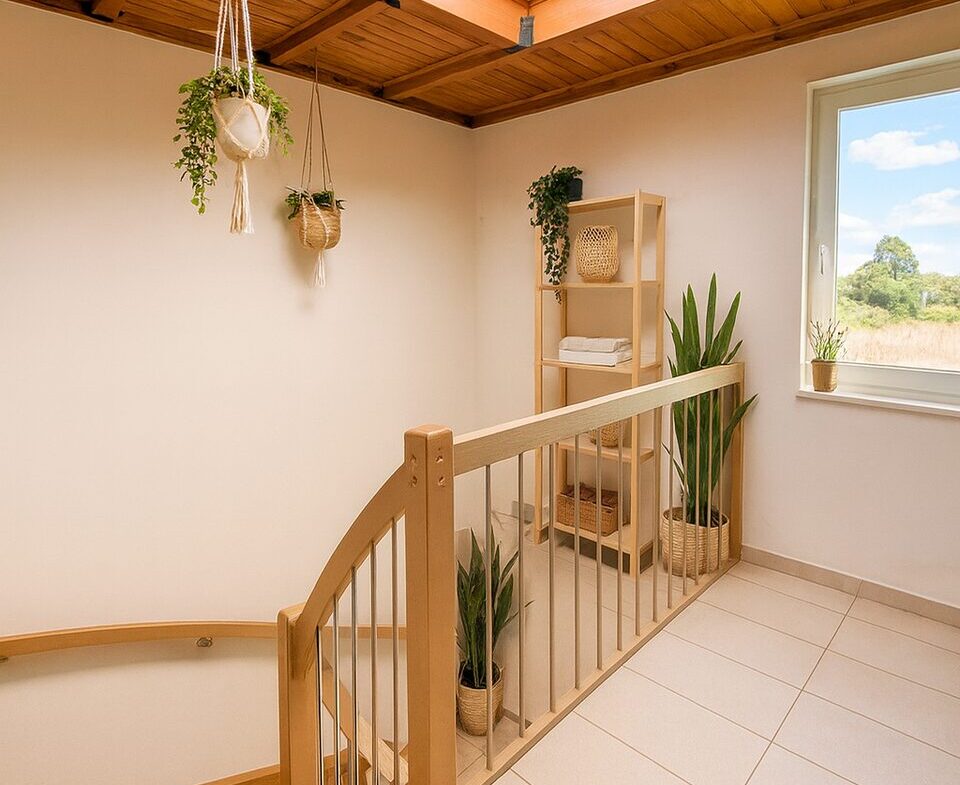 Bright stair landing with exposed wooden beams and a skylight, featuring hanging plants, a wooden railing, and a tall shelf with towels and greenery by a large window showing a outdoor field.