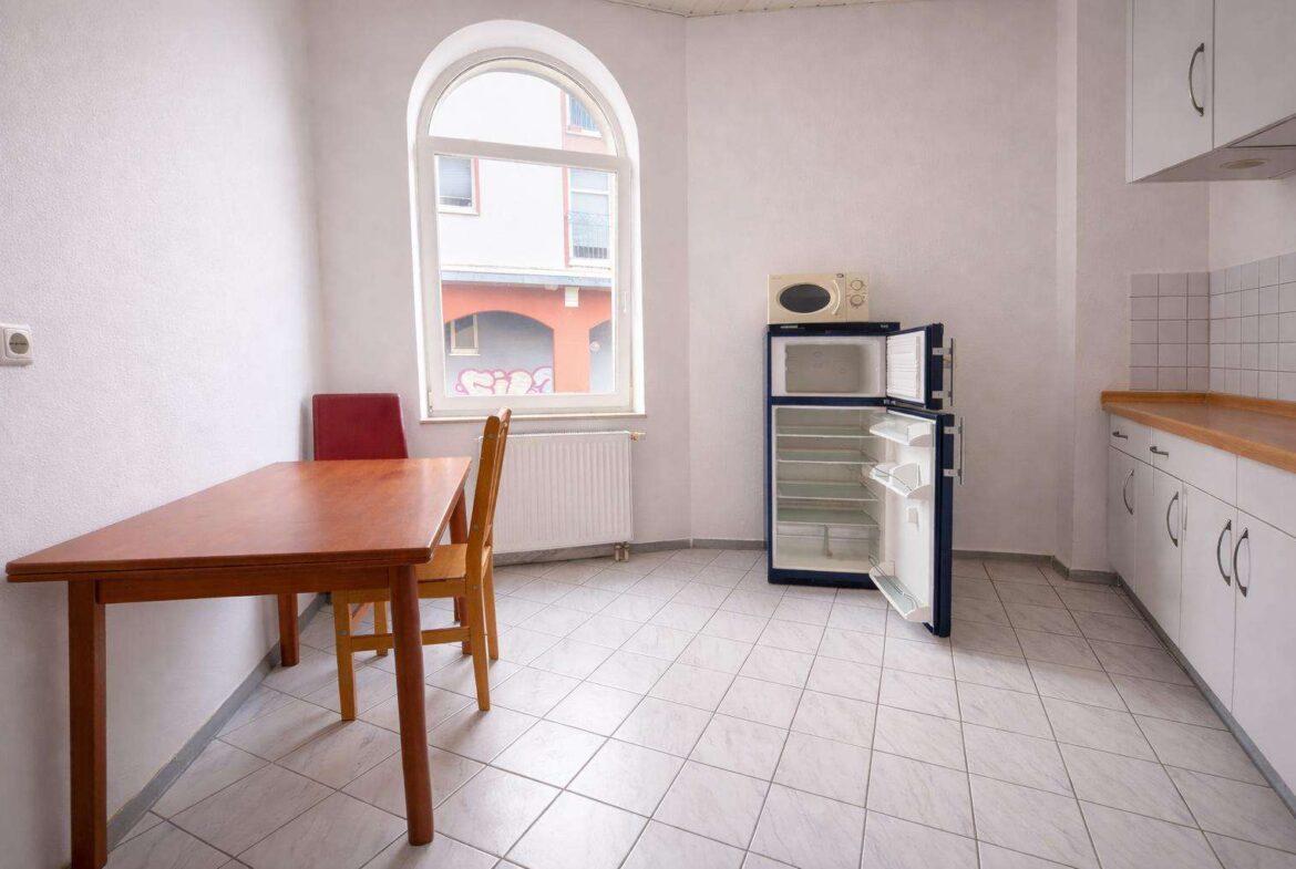 Small kitchen with a wooden table and chairs, an open refrigerator with empty shelves, a microwave on top, white cabinets, and a tiled floor under a large arched window.