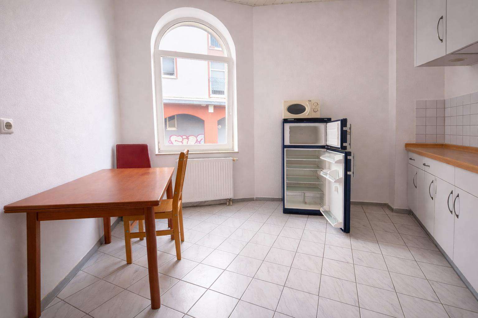 Small kitchen with a wooden table and chairs, an open refrigerator with empty shelves, a microwave on top, white cabinets, and a tiled floor under a large arched window.