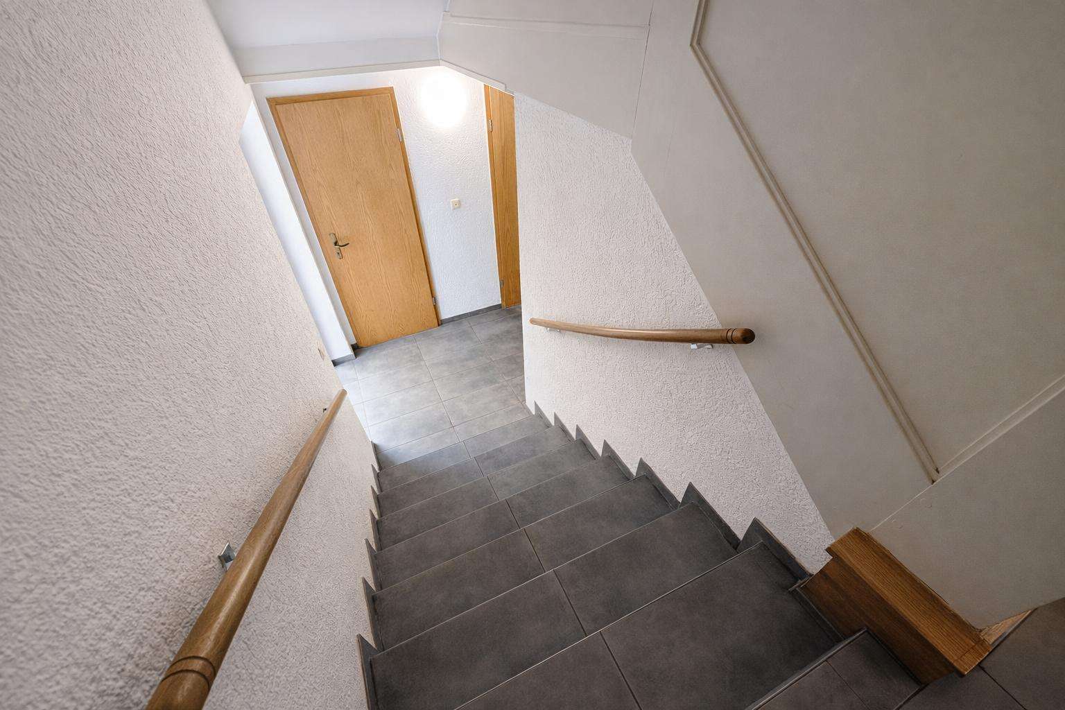 Top-down view of a stairwell with gray tile steps, wooden handrails on both sides, and white textured walls leading to wooden doors at the landing.