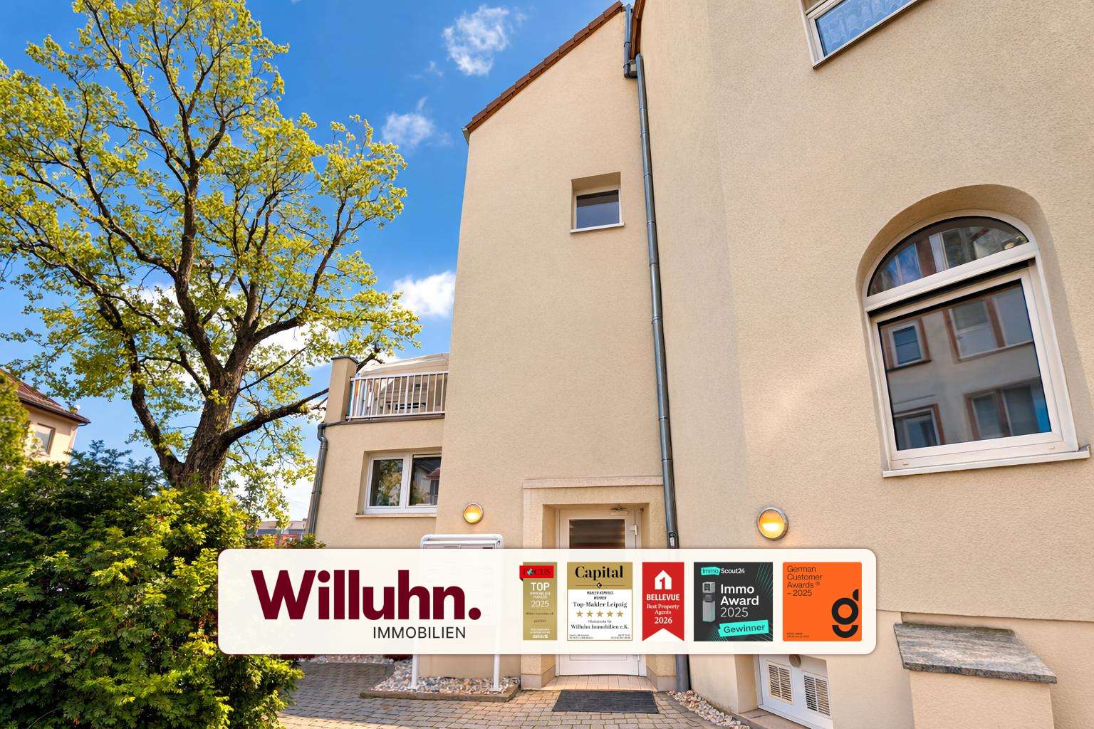 Beige residential building with a large tree on the left and a white Willuhn Immobilien banner with awards across the bottom edge.