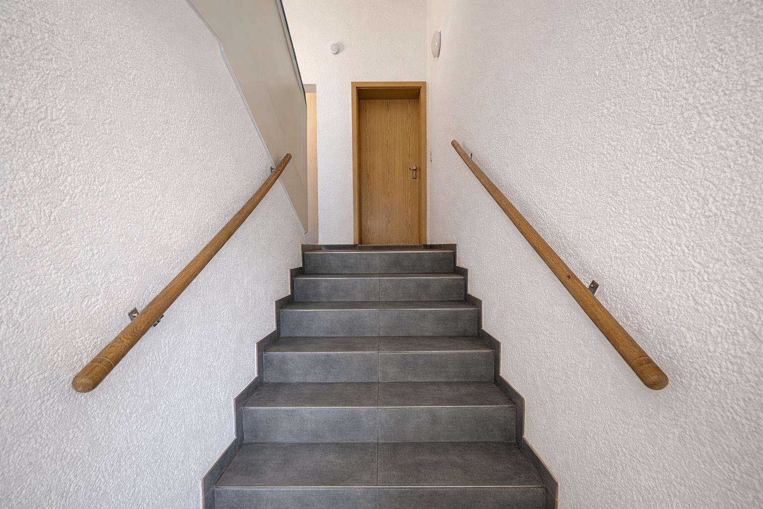 Staircase with grey tiles leads to a wooden door at the top, with wooden handrails on white textured walls.