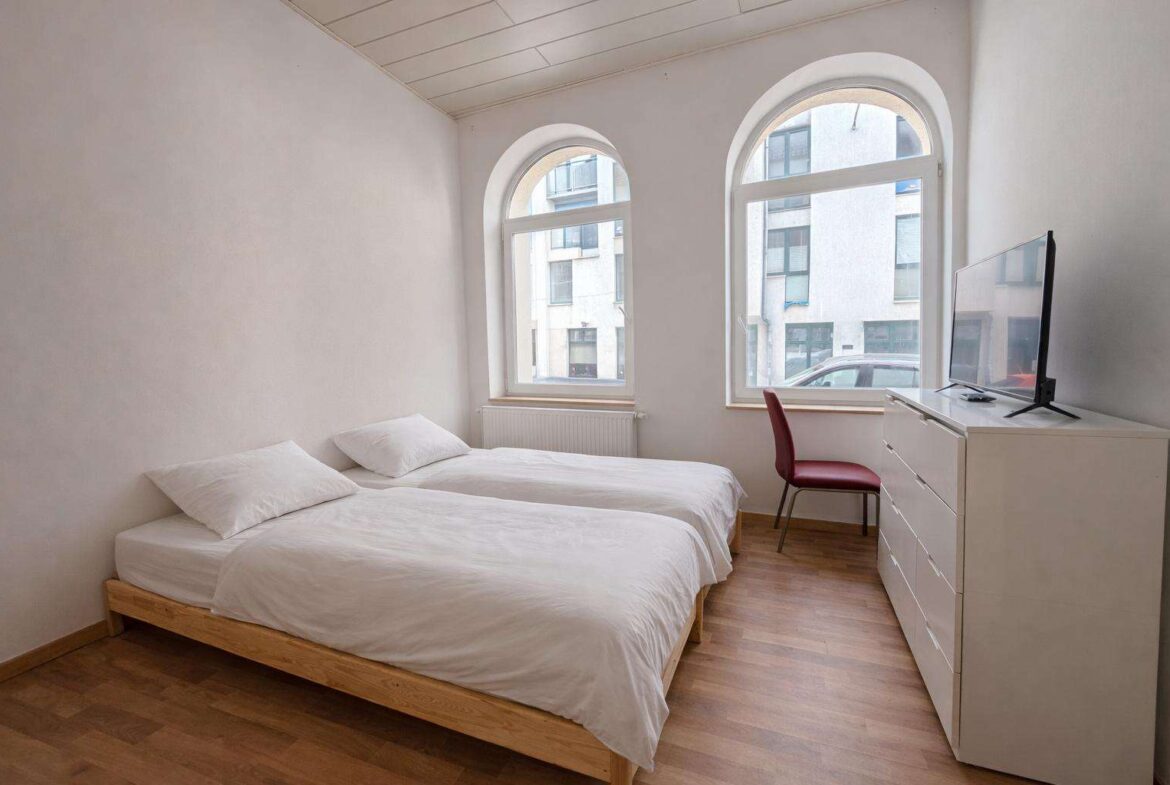 Minimalist twin bedroom with two single beds, white bedding, and wooden frames facing a white dresser with a TV; arched windows on the wall behind.