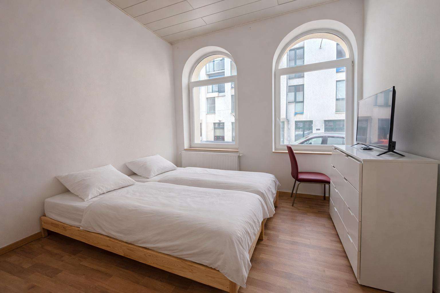 Minimalist twin bedroom with two single beds, white bedding, and wooden frames facing a white dresser with a TV; arched windows on the wall behind.