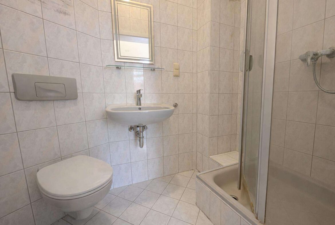 Small tiled bathroom with a wall-hung toilet, oval sink, mirror above, glass shelf, and a corner shower with sliding door.