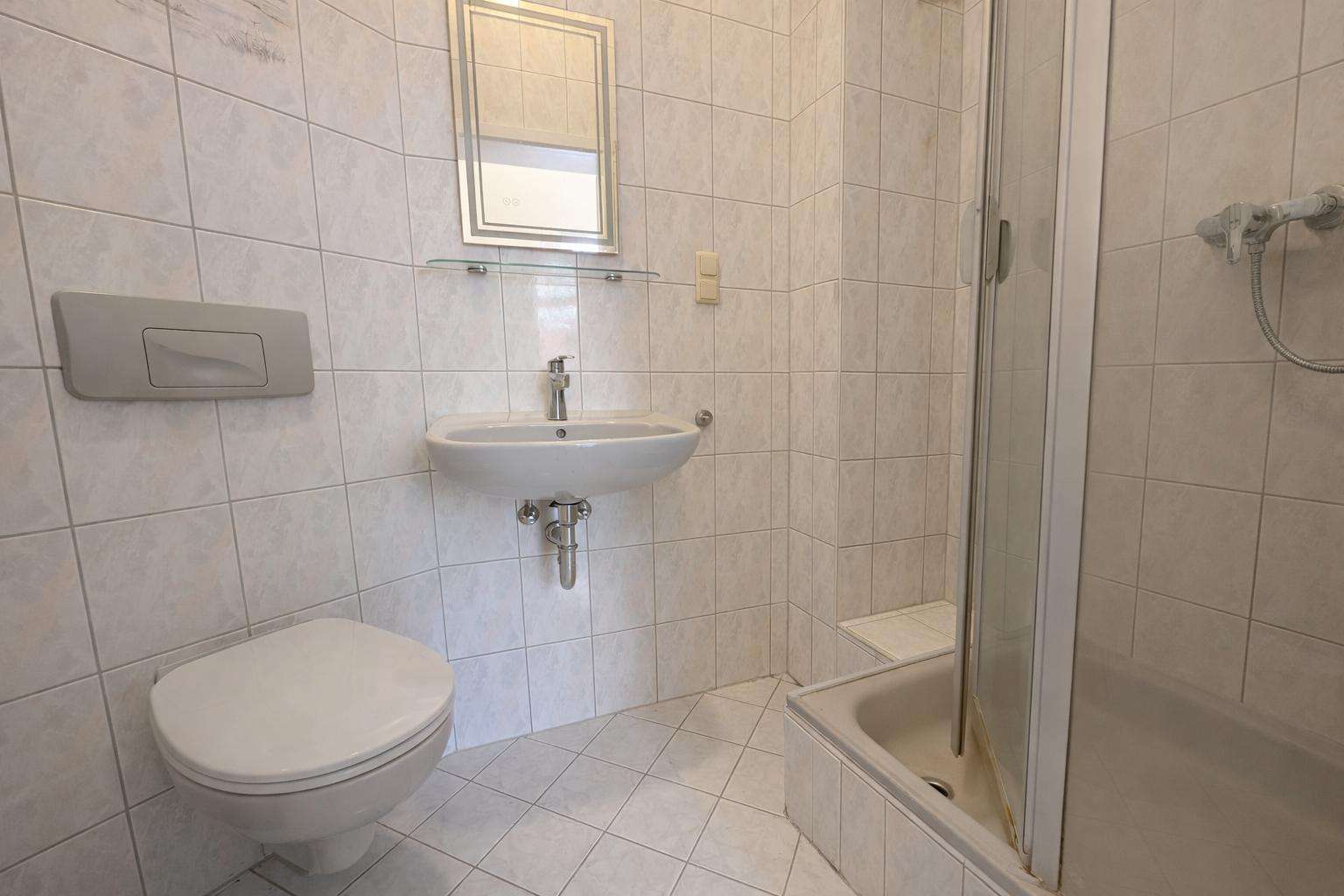Small tiled bathroom with a wall-hung toilet, oval sink, mirror above, glass shelf, and a corner shower with sliding door.