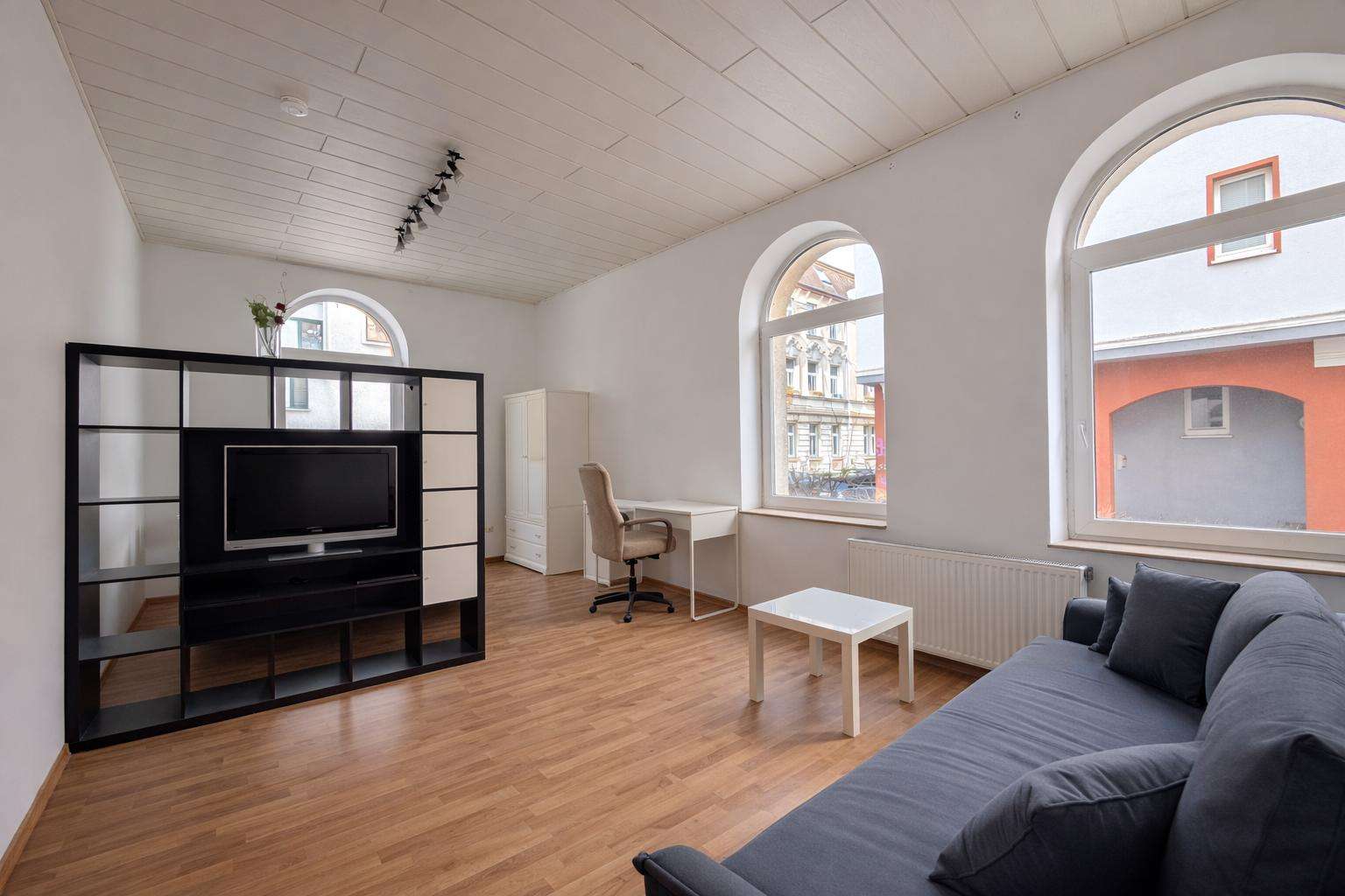 Living room with a black entertainment center, TV, white cabinet, and gray sofa facing large arched windows with wooden floor and a small white coffee table.",