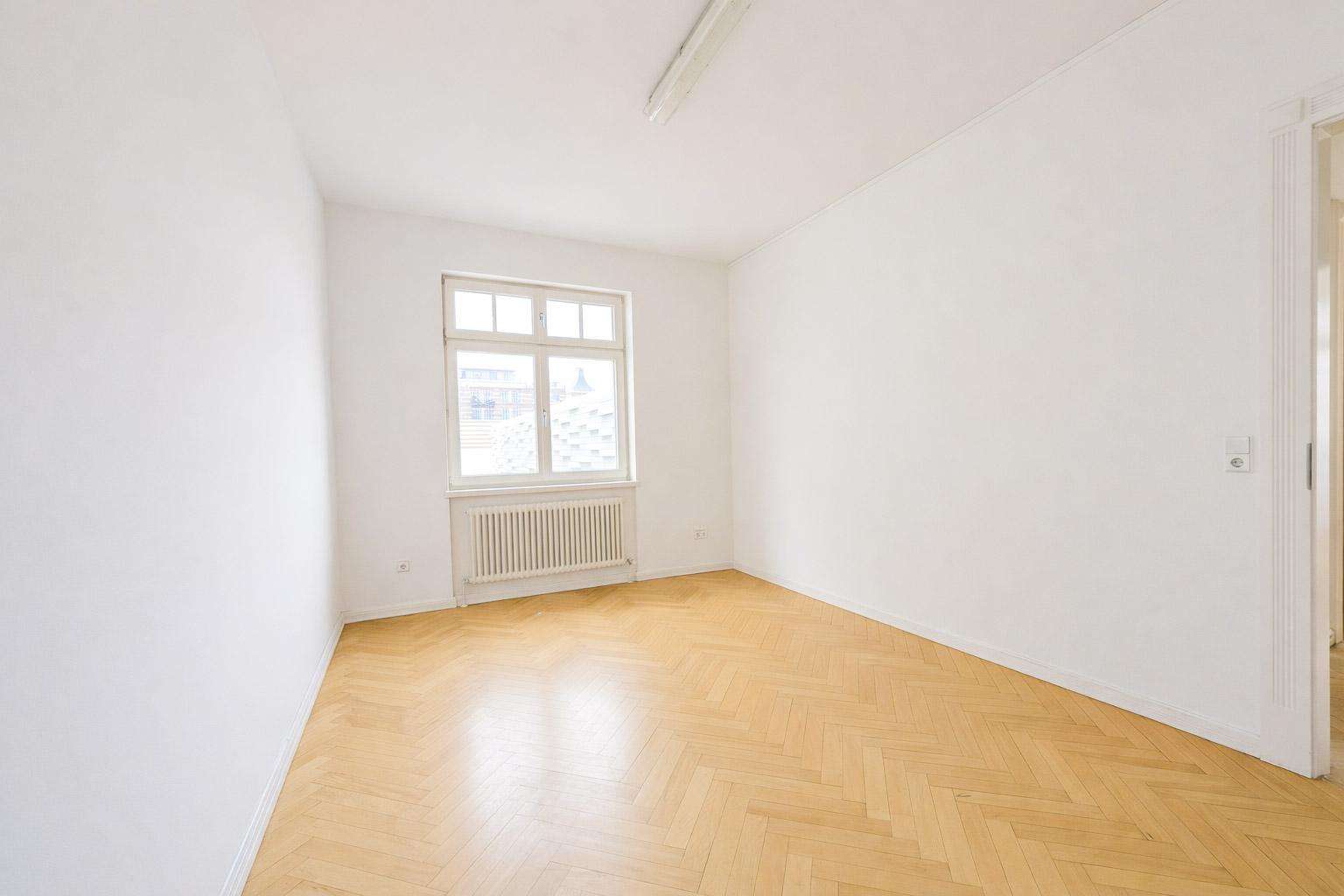 Empty white room with a window, radiator beneath, and light wood parquet floor.