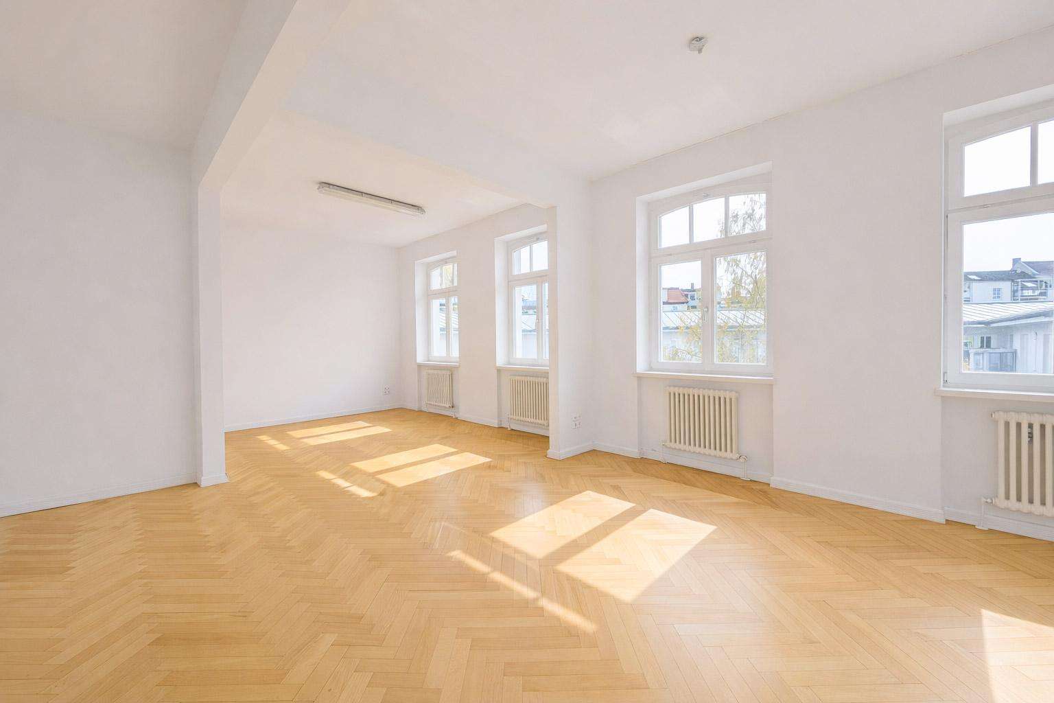 Bright empty room with white walls and a light wood herringbone floor, illuminated by multiple large windows casting sunlight patterns on the floor.