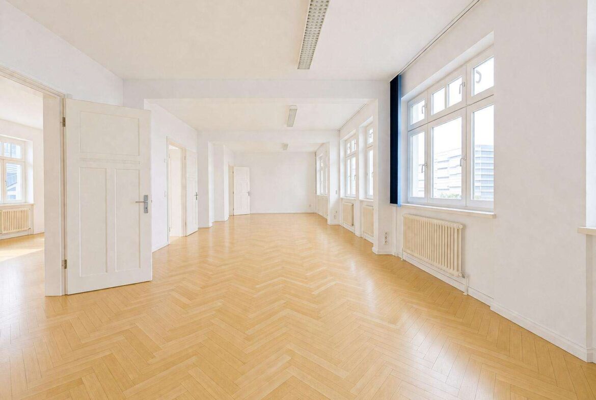 Bright, empty office corridor with white walls, large windows, and light-wood chevron flooring.