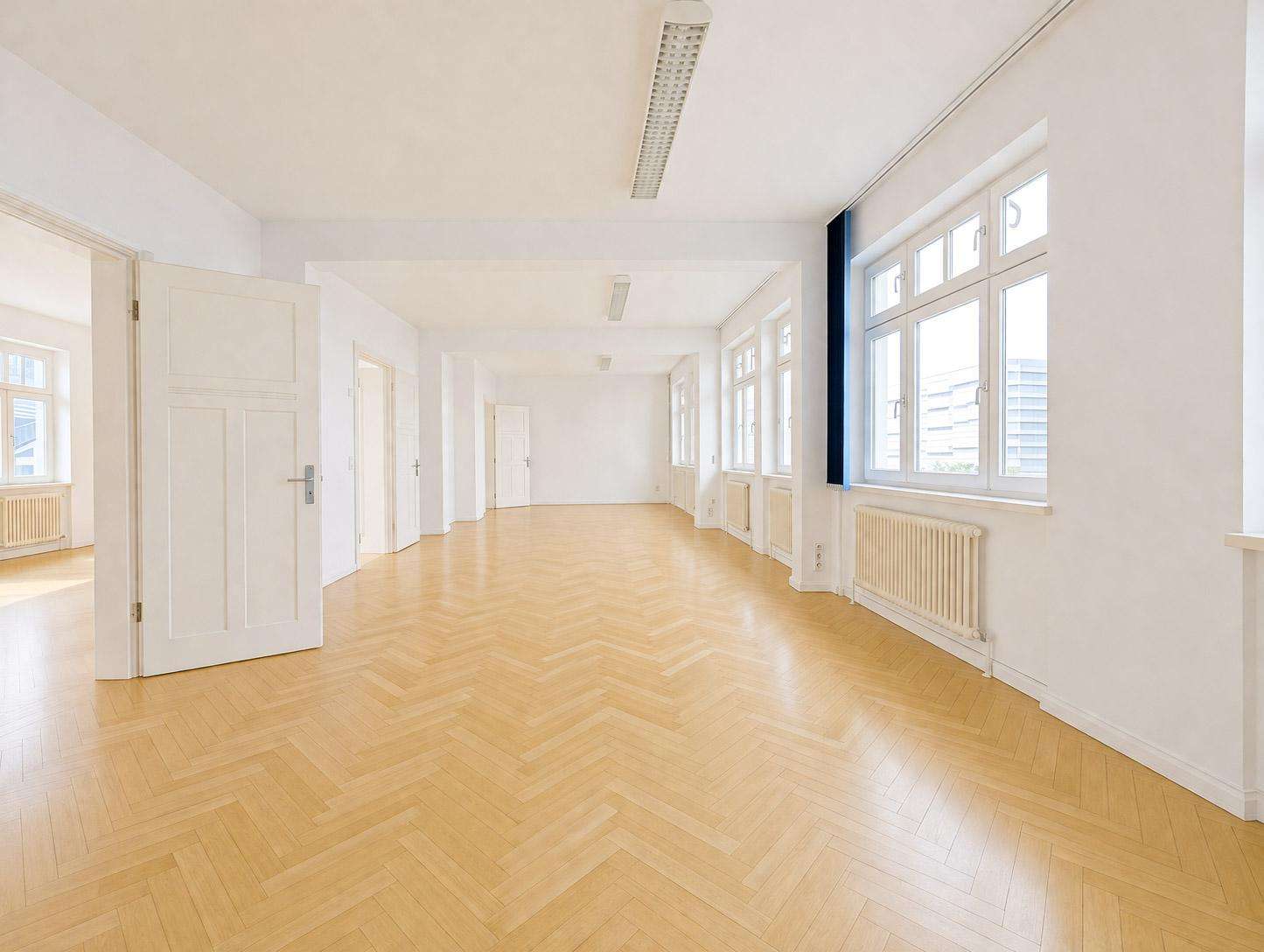 Bright, empty office corridor with white walls, large windows, and light-wood chevron flooring.