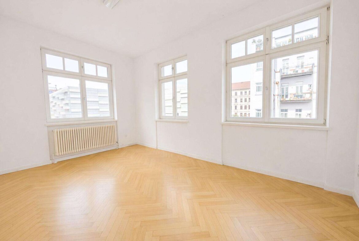Bright empty room with three large windows, white walls, and a light wooden herringbone floor; radiator along one wall
