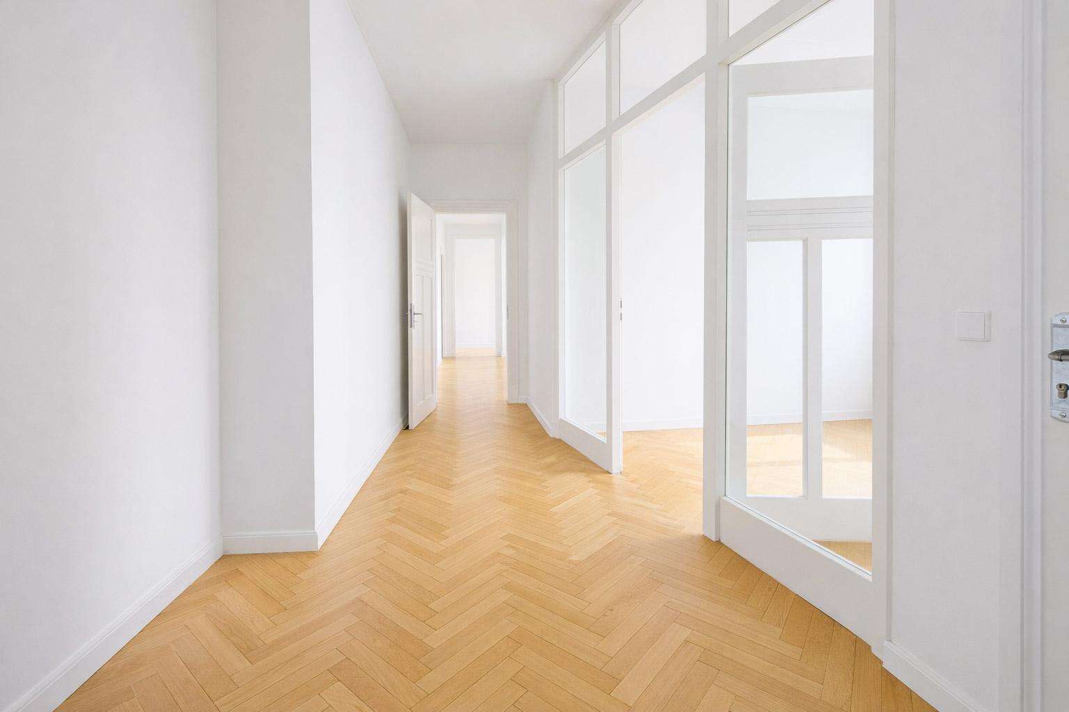 Bright white hallway with tall glass-paneled doors on the right and a light herringbone wooden floor