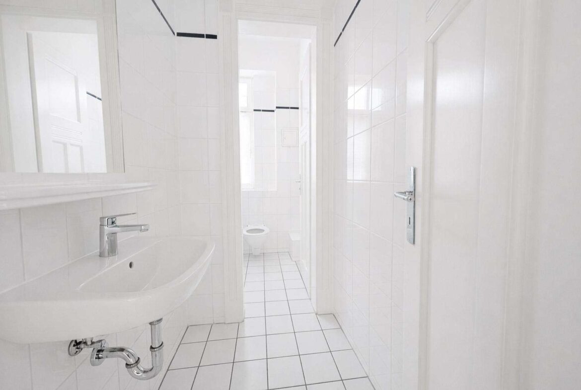 White tiled bathroom with a wall-mounted sink and chrome plumbing, visible through a doorway into a narrow hall leading to a toilet in the background