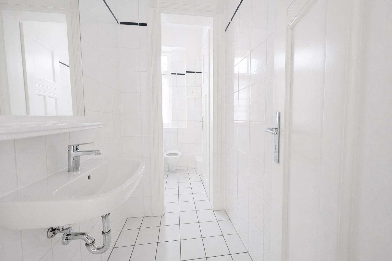 White tiled bathroom with a wall-mounted sink and chrome plumbing, visible through a doorway into a narrow hall leading to a toilet in the background
