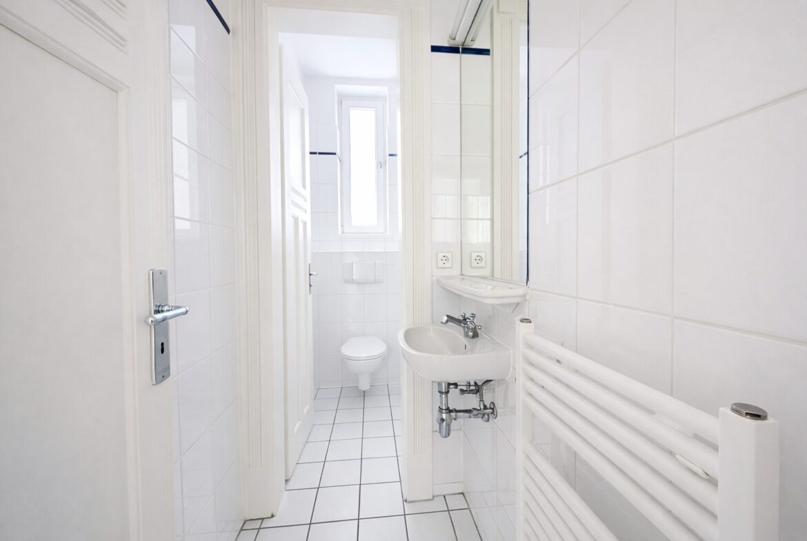 Small bright white bathroom with wall-mounted sink, toilet, radiator, and a tall window in back.