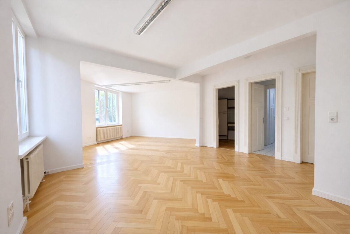 Bright empty living area with herringbone wood floor, large windows, and white walls; radiator under the window.