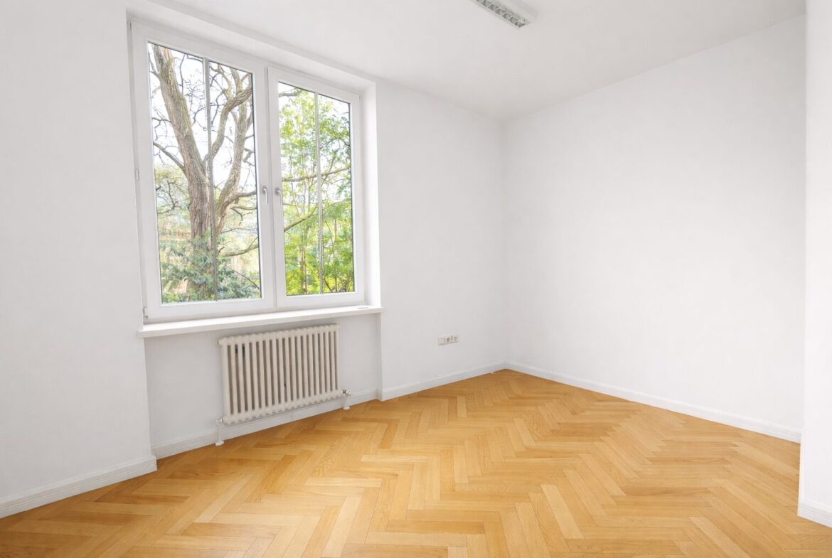 Bright empty room with large window overlooking trees, radiator under window, and herringbone wooden floor