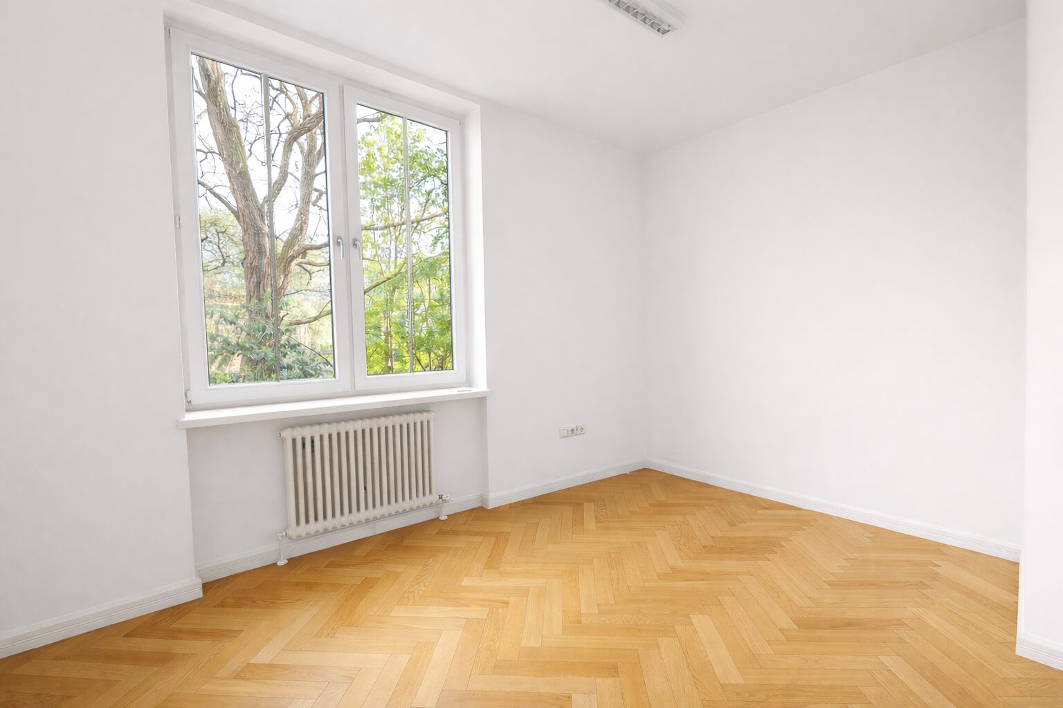 Bright empty room with large window overlooking trees, radiator under window, and herringbone wooden floor