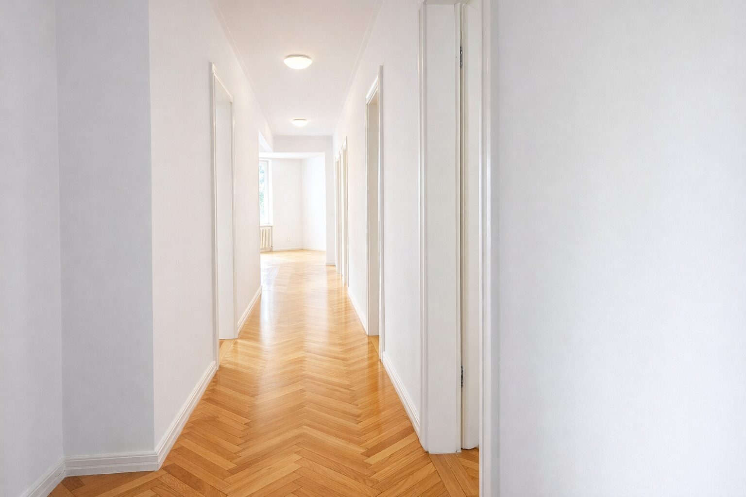 Bright white hallway with chevron hardwood floor and multiple doors along both sides, leading to a sunlit room at the end.