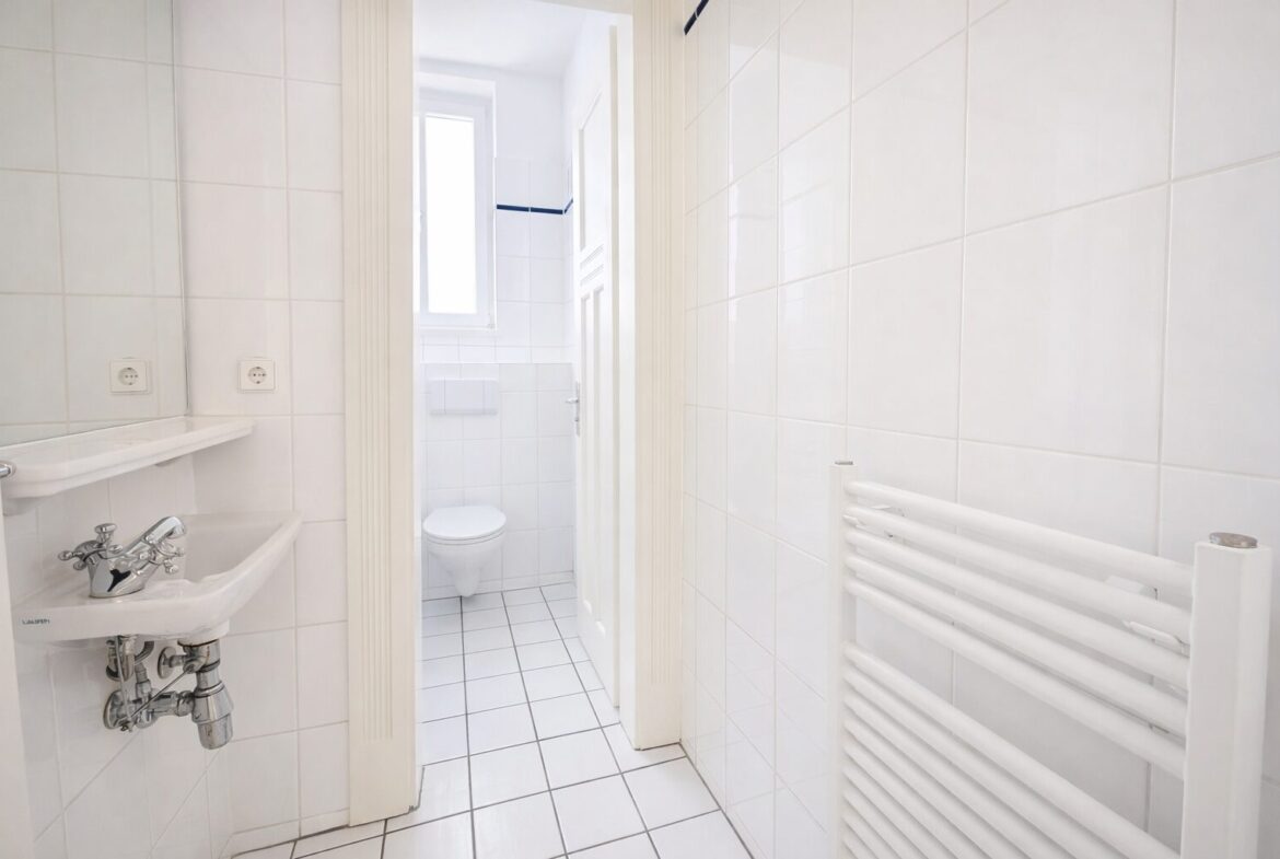 Small white bathroom with tiled walls and floor, sink on left with exposed plumbing, and a toilet at the far end under a window; a white towel radiator on the right.