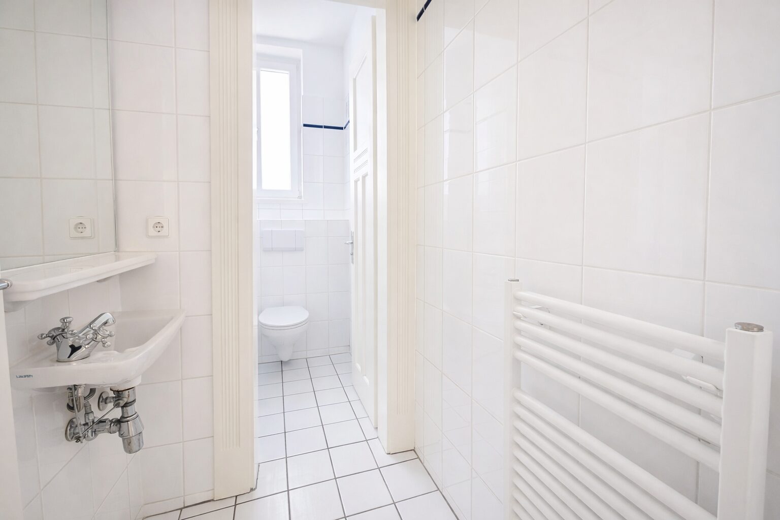 Small white bathroom with tiled walls and floor, sink on left with exposed plumbing, and a toilet at the far end under a window; a white towel radiator on the right.