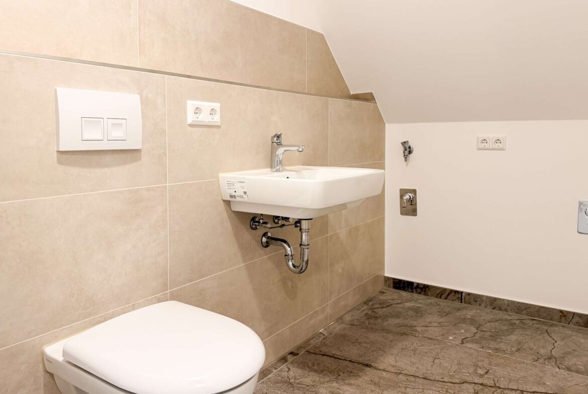 Modern bathroom corner with a white wall-mounted sink, beige tiled wall, and a white toilet in the foreground.