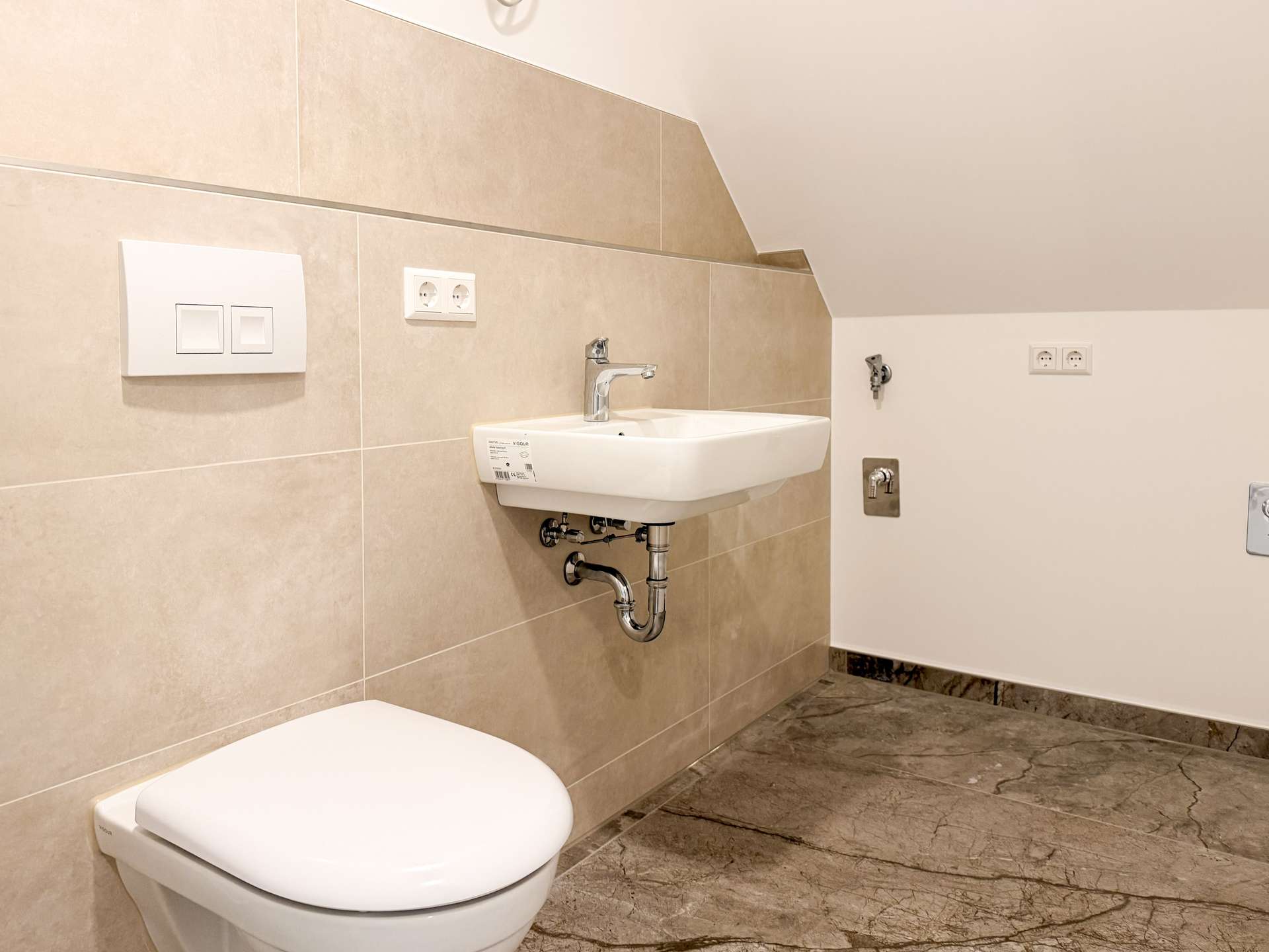 Modern bathroom corner with a white wall-mounted sink, beige tiled wall, and a white toilet in the foreground.
