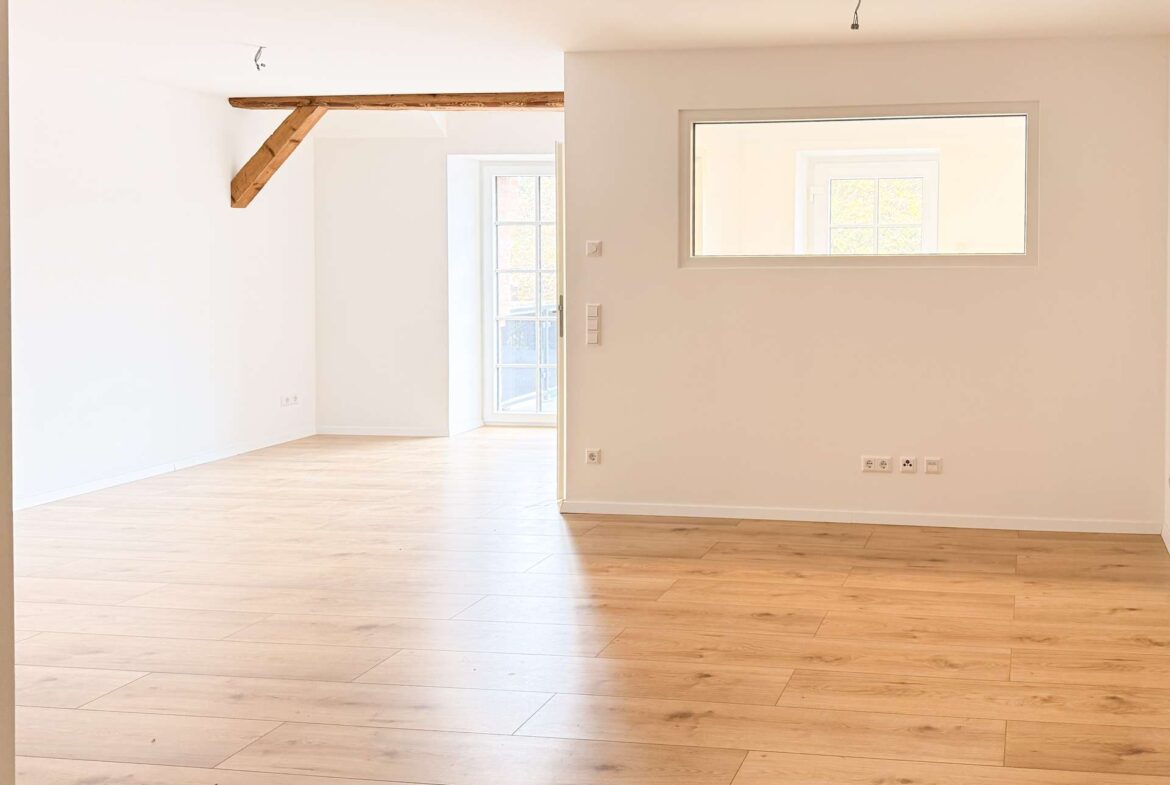 Bright empty room with light wood floors, white walls, and exposed wooden beams; doorway to a balcony and a rectangular window on the wall.