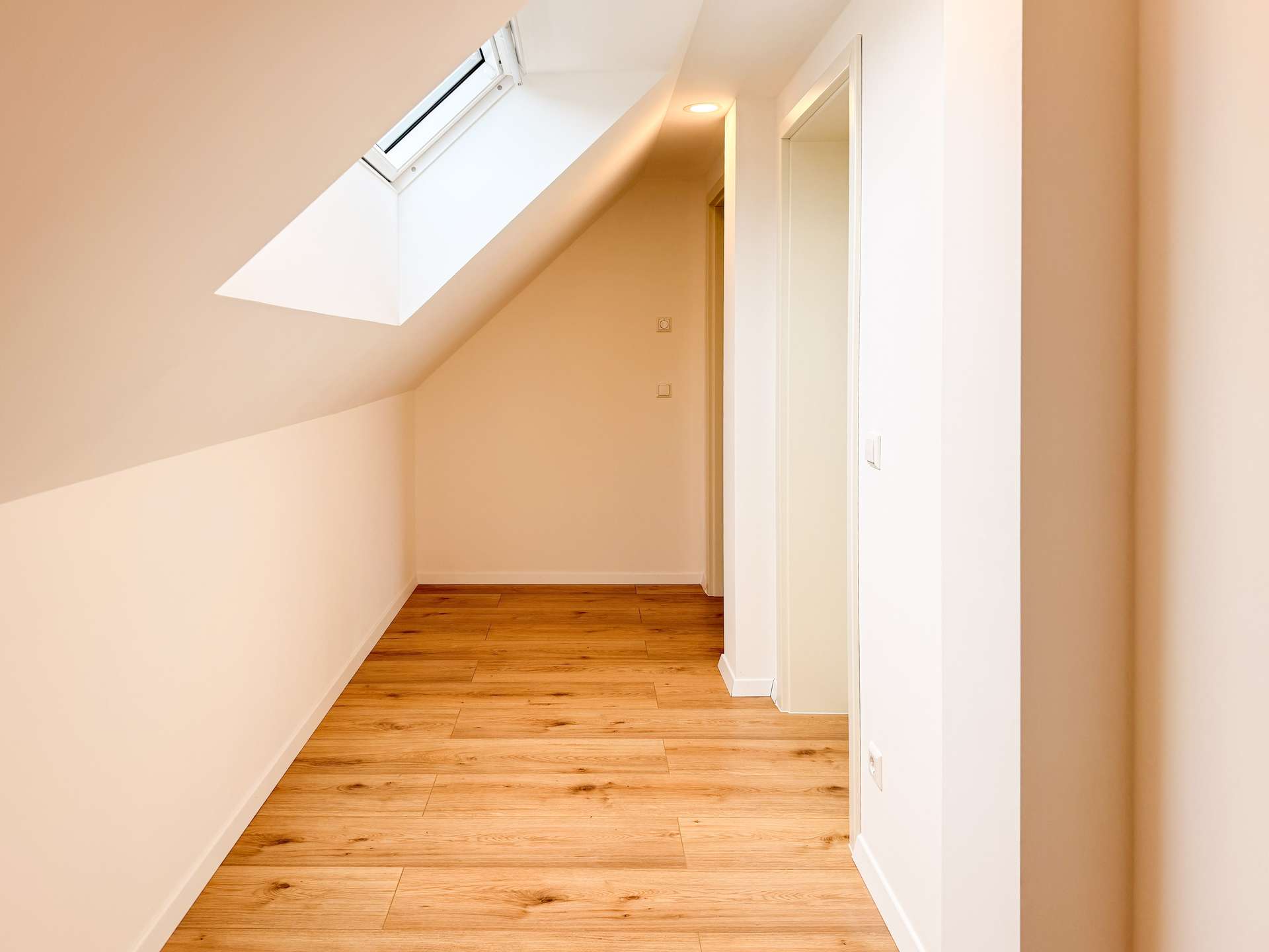 Bright narrow hallway with a sloped ceiling and skylight, white walls and warm wooden flooring there