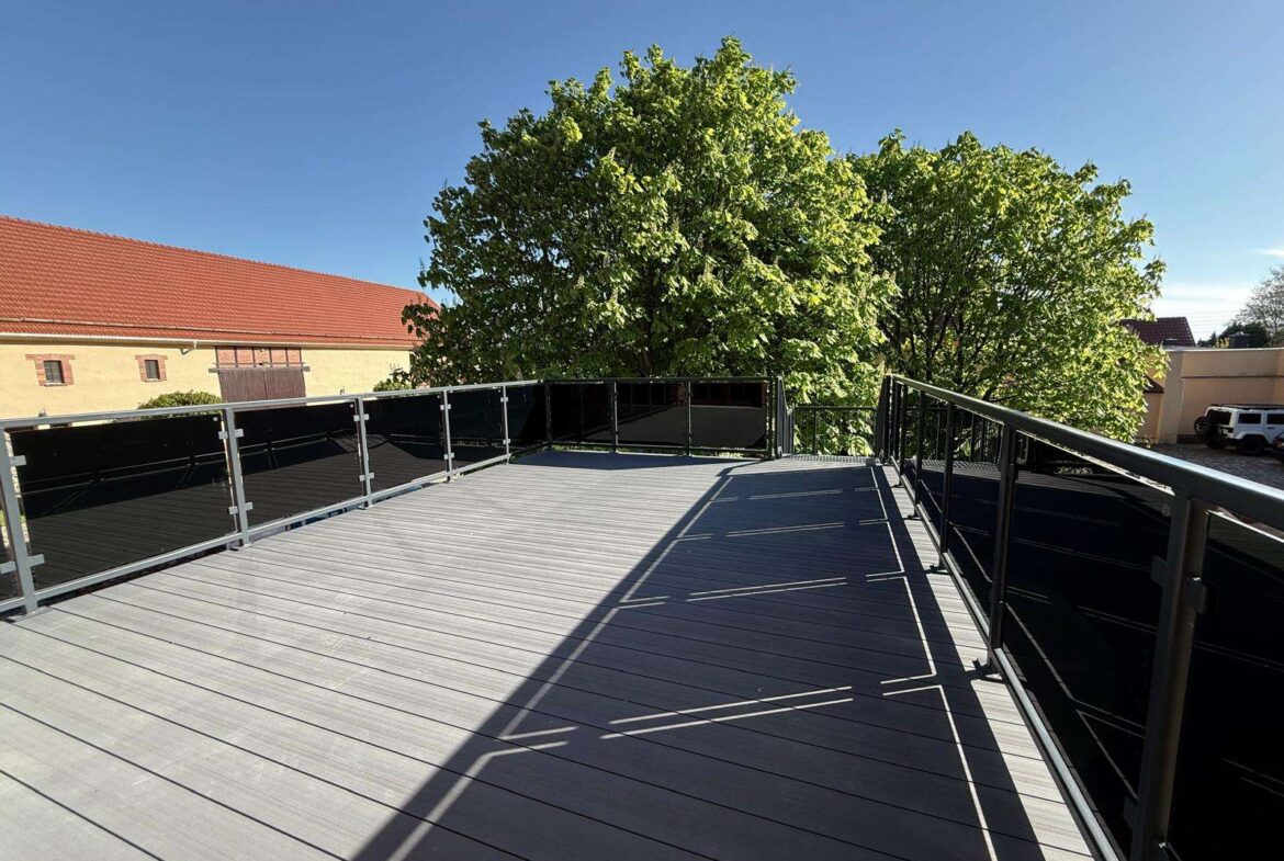 Rooftop terrace with metal-and-glass railing, gray wooden decking, and large green trees beyond under a clear blue sky.